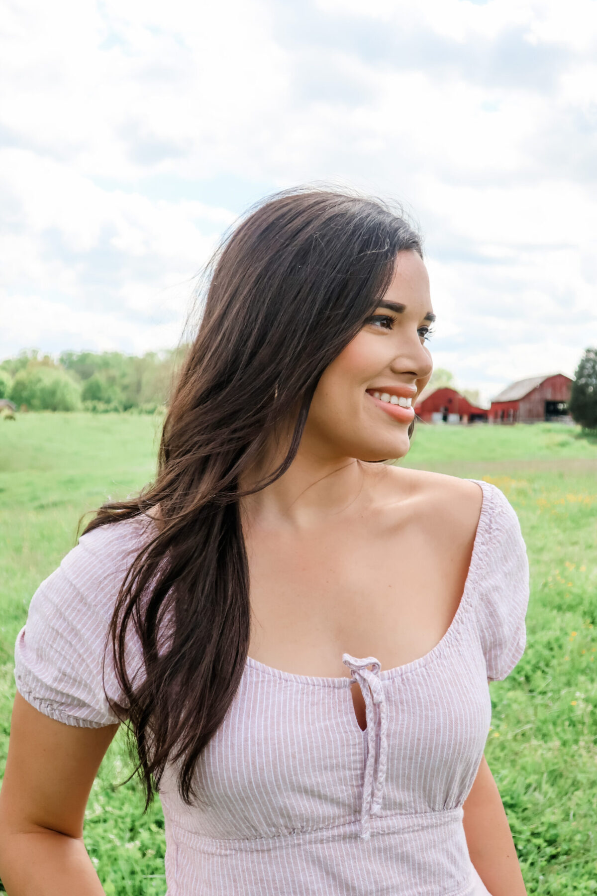 A woman with long dark hair standing in a field, smiling and looking away from the camera, into the distance