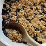 a close up view of a dish of einkorn blueberry crisp, with a wooden spoon scooping out a portion