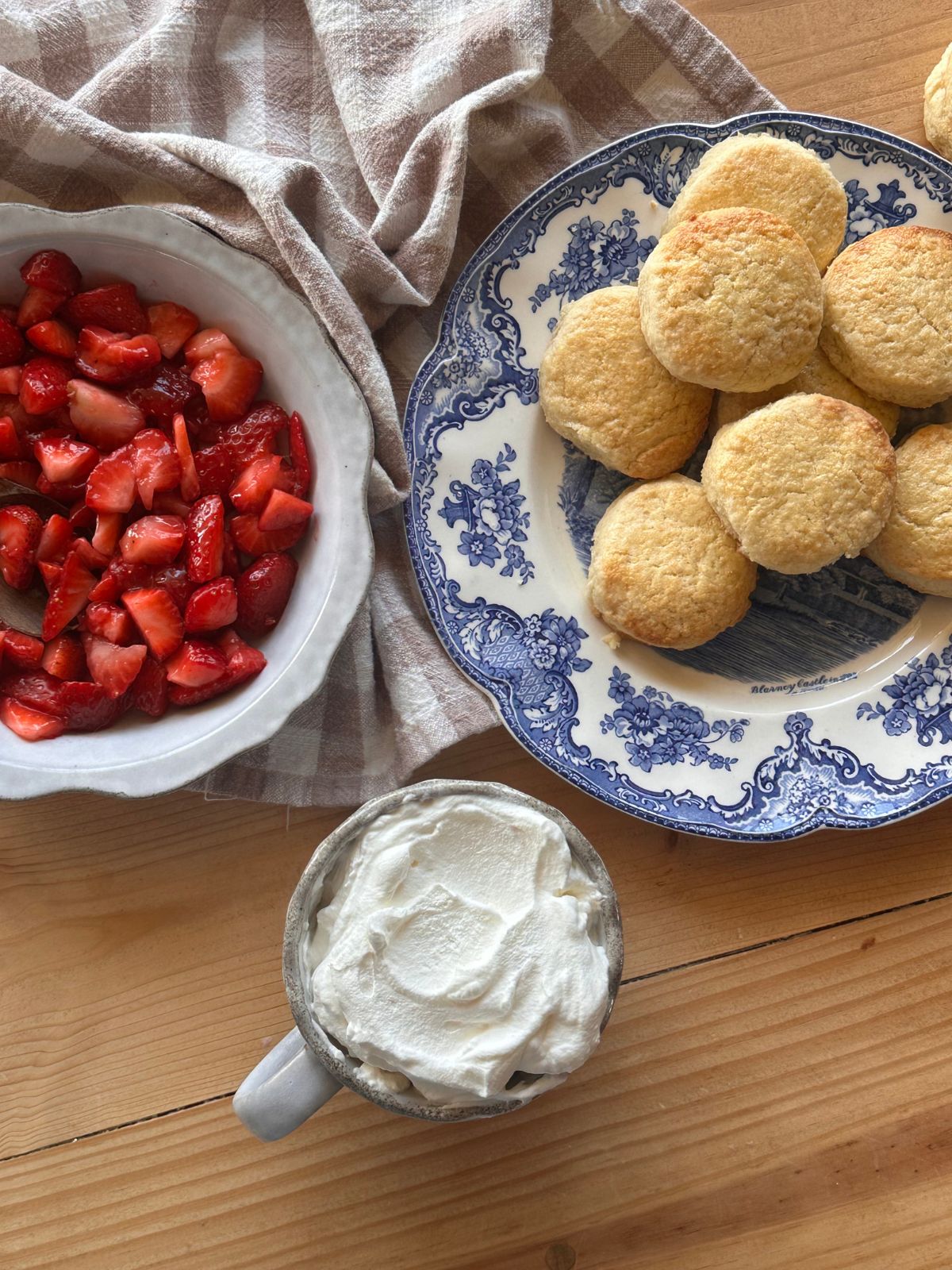 a plate of biscuits, a cup of whipped cream, and a bowl of chopped strawberries are arranged on a wooden countertop