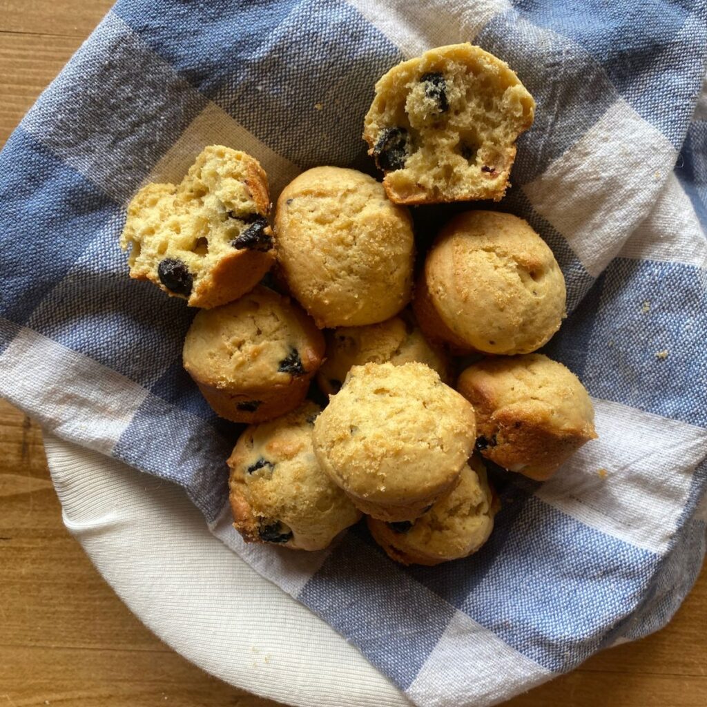 A basket of golden-brown einkorn blueberry muffins nestled in a blue and white checkered cloth, with one muffin split open to reveal the fluffy, berry-filled interior.