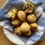 A basket of golden-brown einkorn blueberry muffins nestled in a blue and white checkered cloth, with one muffin split open to reveal the fluffy, berry-filled interior.