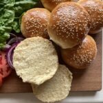 burger buns with sesame seeds on top laying on a wooden cutting board with lettuce and tomato