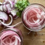 two small glass jars full of slicked pickled red onions sitting on a wooden cutting board