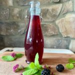 A glass bottle filled with blackberry simple syrup sits on a wooden board, surrounded by fresh mint leaves, blackberries, and small pink flowers, set against a stone wall.