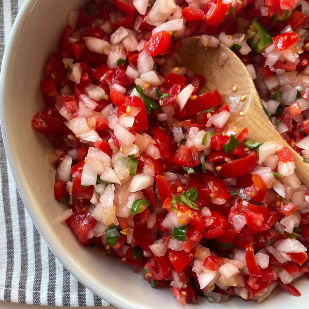 Close-up of a vibrant salsa in a bowl, featuring chopped red tomatoes, white onions, green onion, and a wooden spoon, on a striped cloth background.