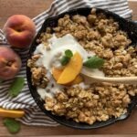 peach blueberry crisp in a small cast iron skillet laying on a wooden cutting board with ice cream and fresh peaches on top