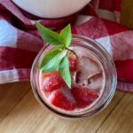 Glass of refreshing strawberry basil lemonade with ice cubes and fresh basil garnish, set on a rustic wooden table with a red and white checkered cloth in the background.