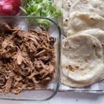 shredded beef in a glass dish with homemade tortillas and taco toppings