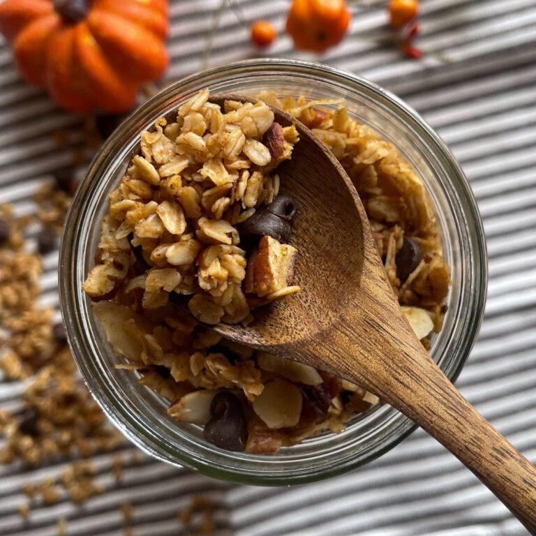 Overhead view of homemade pumpkin spice granola in a glass jar, filled with oats, chocolate chips, nuts, and toasted coconut, set on a striped cloth with fall decor in the background.