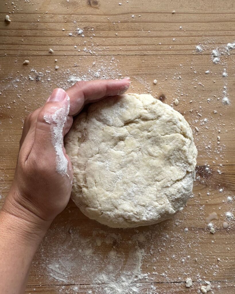 a small disc of sourdough pie crust on a wooden surface. A hand is cupped around the outside edge of the disc