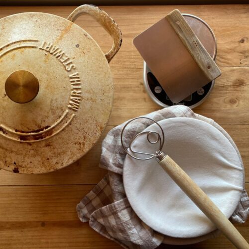 Essential sourdough bread baking tools including a Dutch oven, bench scraper, digital scale, Danish dough whisk, and proofing bowl on a wooden countertop.