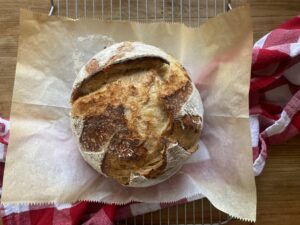 a round baked loaf of sourdough bread laying on a piece of parchment paper with a red and white checkered towel
