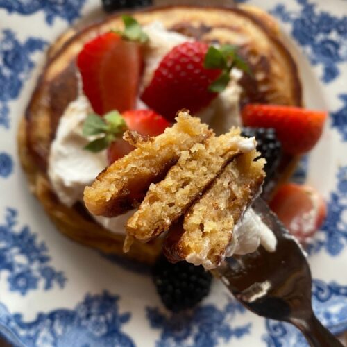 a stack of fluffy pancakes made with fresh milled flour on a blue and white plate and topped with whipped cream and fruit