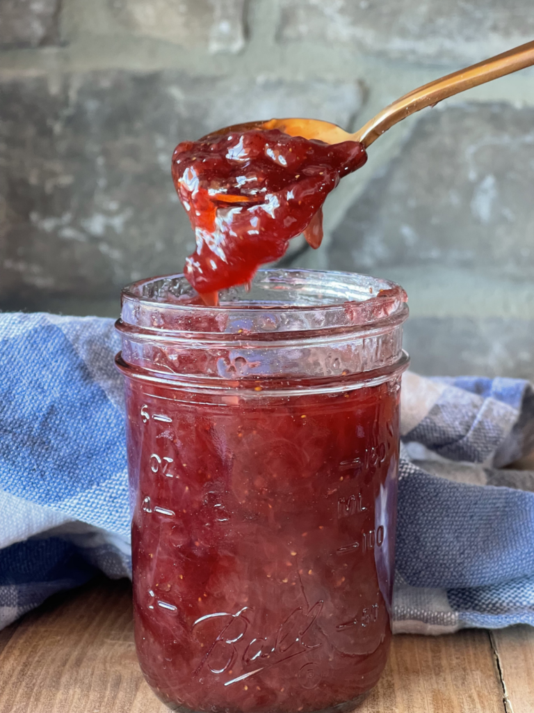 a small glass jar full of homemade strawberry jam. A spoon is hovering above the jar with a scoop of jam slowly falling down into the jar