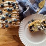 a loaf of sourdough lemon blueberry quick bread on a wooden countertop next to a slice of bread on a white plate