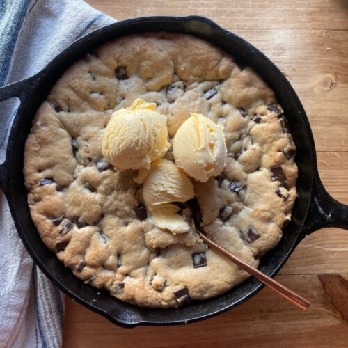 A large, golden-brown chocolate chip cookie baked in a black cast-iron skillet, topped with three scoops of vanilla ice cream. A spoon is placed in the cookie, and a white and blue dish towel is partially visible on the side.
