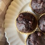 Close-up of three chocolate muffins in beige paper liners on a scalloped white plate, with a checked cloth beside them.
