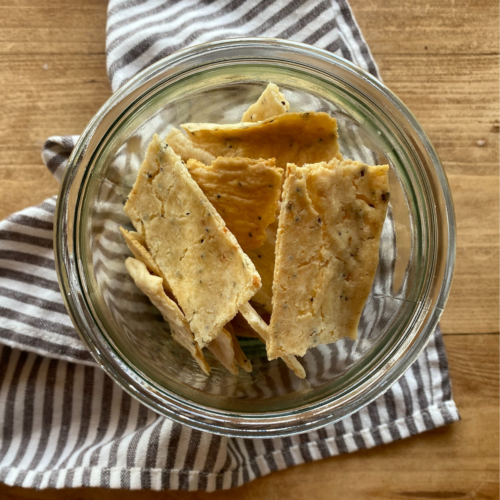 Top-down view of rectangular sourdough discard crackers in a glass jar on a wooden surface. A striped cloth is underneath, adding a rustic, cozy feel.