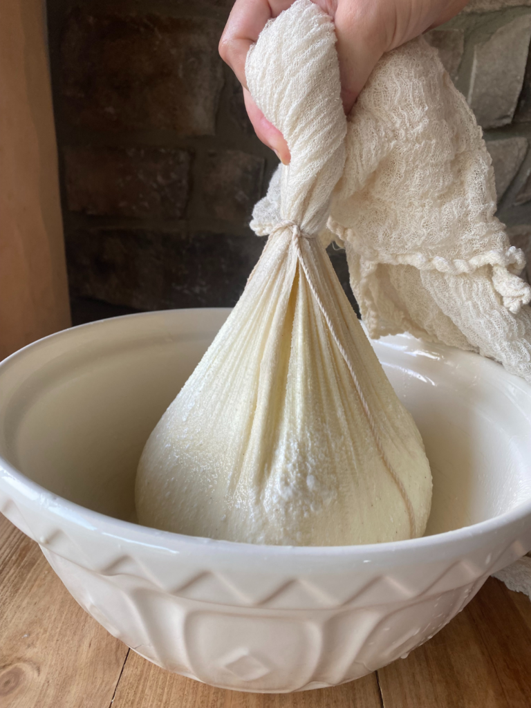 Straining homemade Greek yogurt in cheesecloth over a ceramic bowl to remove whey and thicken the texture.