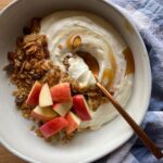 Bowl of Greek yogurt topped with apple slices, granola, and a drizzle of honey, with a copper spoon and blue kitchen towel in the background.