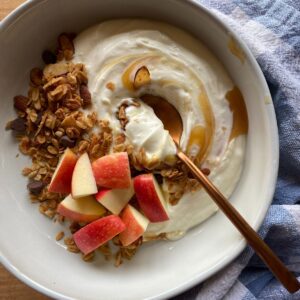 Bowl of Greek yogurt topped with apple slices, granola, and a drizzle of honey, with a copper spoon and blue kitchen towel in the background.