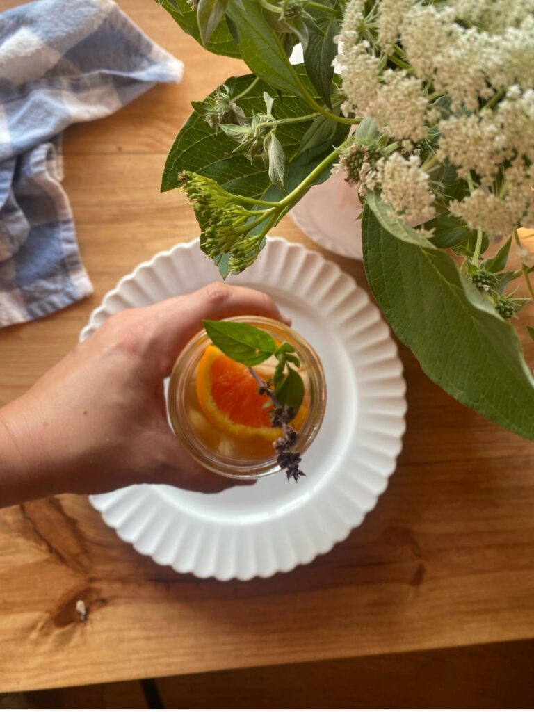 Hand holding a glass of orange iced tea garnished with a fresh orange slice, basil, and lavender, over a white plate on a wooden table with summer florals and a checkered cloth.
