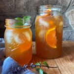 Two large mason jars filled to the top with orange iced tea, sitting on a rustic wooden table. Orange sliced and fresh mint is used as a garnish.
