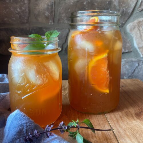 Two large mason jars filled to the top with orange iced tea, sitting on a rustic wooden table. Orange sliced and fresh mint is used as a garnish.