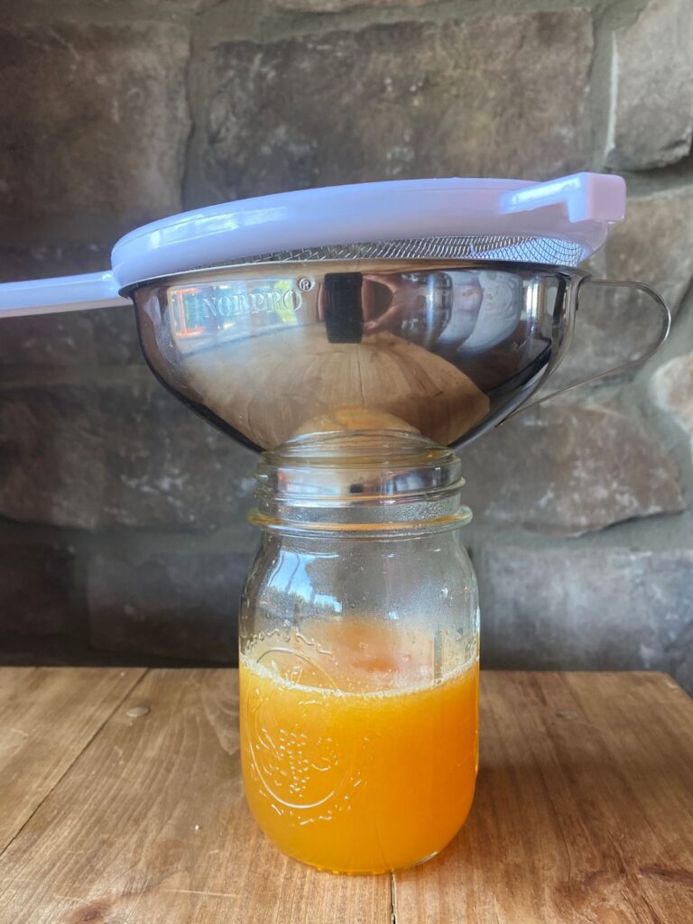 A mason jar filled with fresh orange juice sits on a wooden table. A metal strainer rests on top, against a stone wall.