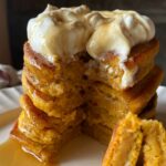 a close up view of a stack of sourdough pumpkin pancakes topped with whipped cream and drizzled with maple syrup