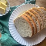 Sliced sourdough sandwich bread on a white plate atop a rustic wooden table, with butter on the side. A green cloth adds contrast, creating a cozy, homemade feel.