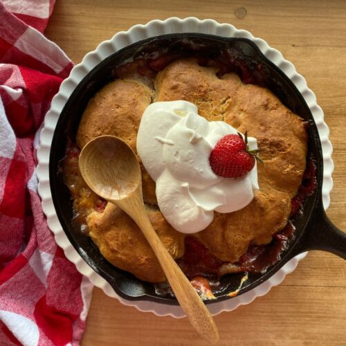 Homemade sourdough strawberry peach cobbler baked in a cast iron skillet, topped with whipped cream and a fresh strawberry, served with a wooden spoon on a rustic wooden table.