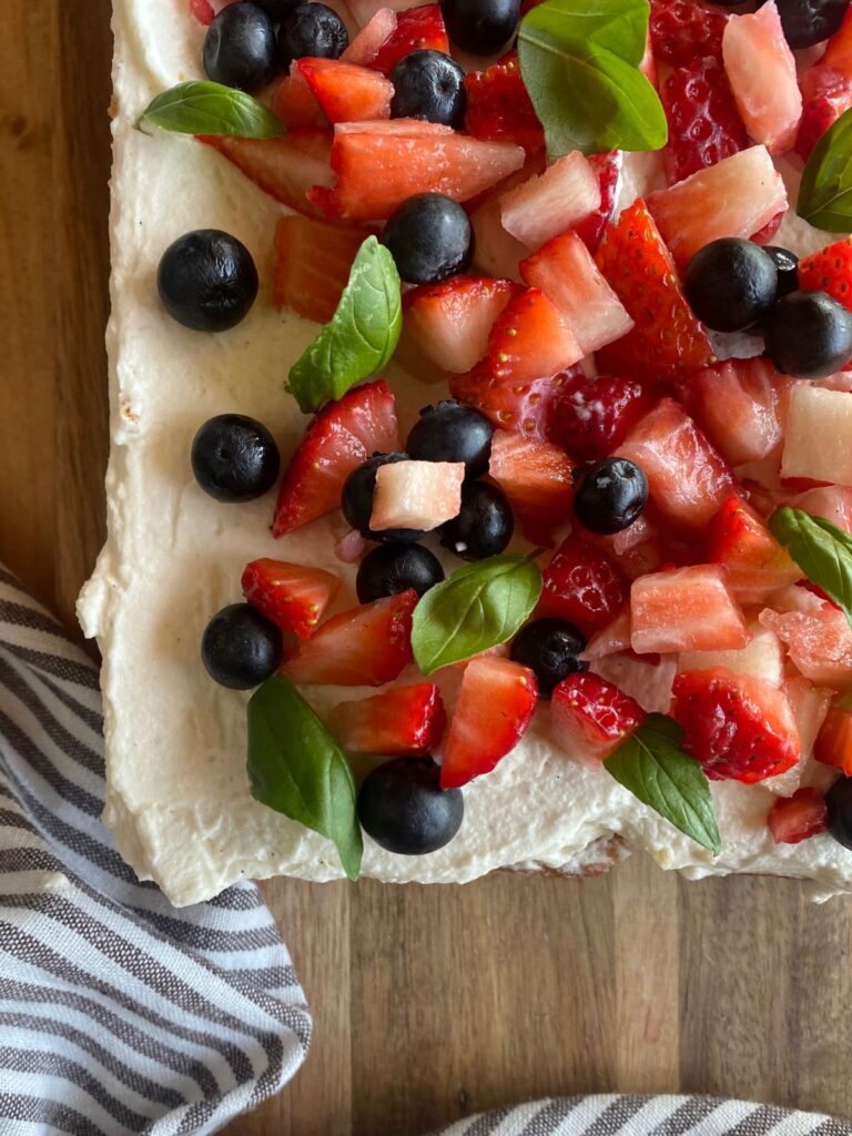 Homemade sourdough vanilla cake topped with fresh strawberries, blueberries, edible flowers, and basil leaves on a rustic wooden surface.