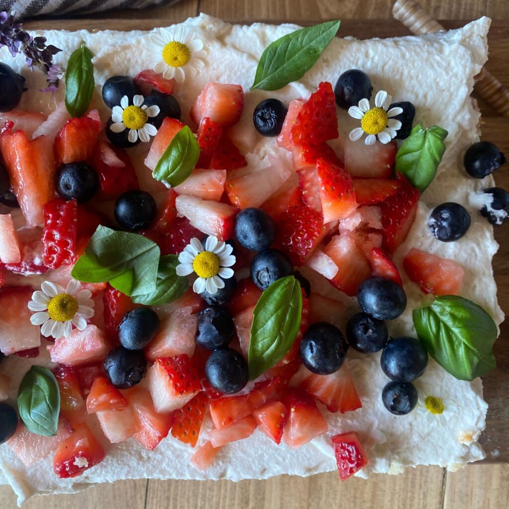 Homemade sourdough vanilla cake topped with fresh strawberries, blueberries, edible flowers, and basil leaves on a rustic wooden surface.