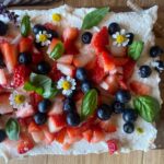 Homemade sourdough vanilla cake topped with fresh strawberries, blueberries, edible flowers, and basil leaves on a rustic wooden surface.
