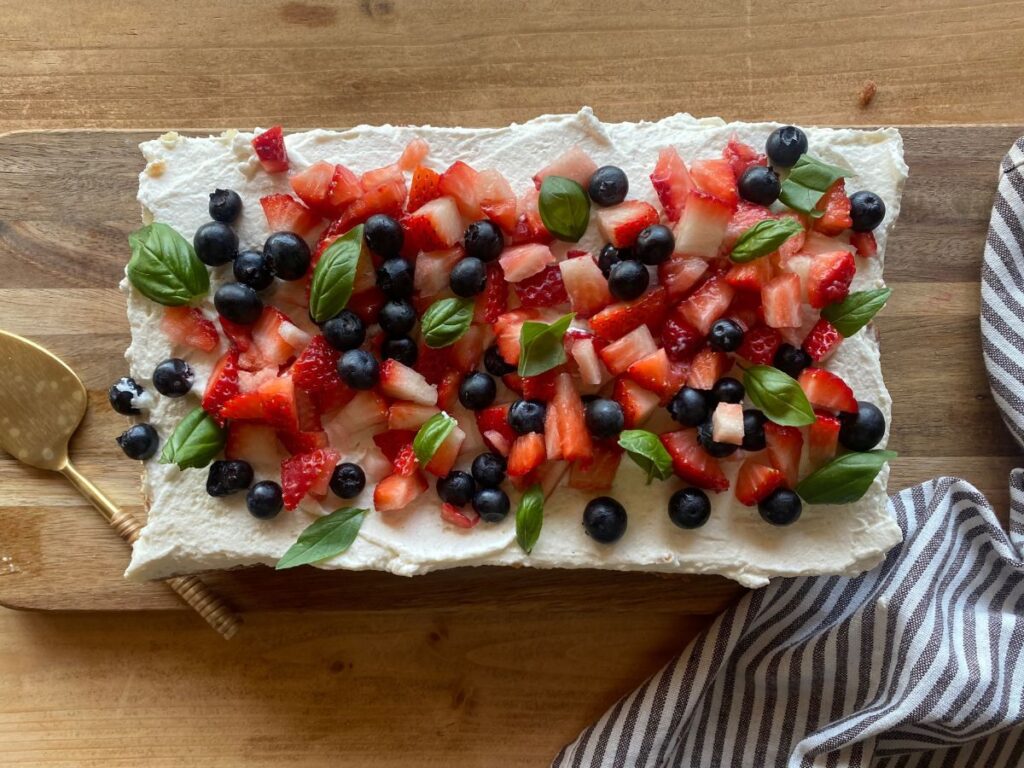 Homemade sourdough vanilla cake topped with fresh strawberries, blueberries, and basil leaves on a rustic wooden surface.