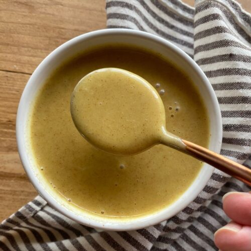 a bowl of homemade honey mustard with a spoon dipped in the middle. A striped cloth lays in the background on a wooden table