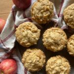 Homemade sourdough apple muffins with a crumb topping, displayed on a dark plate with fresh apples and a red plaid kitchen towel.