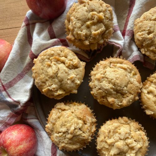 Homemade sourdough apple muffins with a crumb topping, displayed on a dark plate with fresh apples and a red plaid kitchen towel.