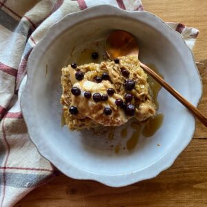 a slice of banana bread baked oatmeal in a gray dish, topped with a drizzle of maple syrup, yogurt, and fresh blueberries. A striped towel lays in the background