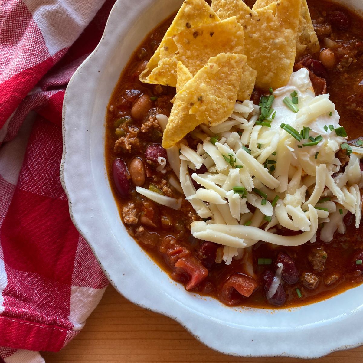 a bowl of hearty chili topped with shredded cheese, tortilla chips, and green onion. A red and white checkered towel lies in the background.