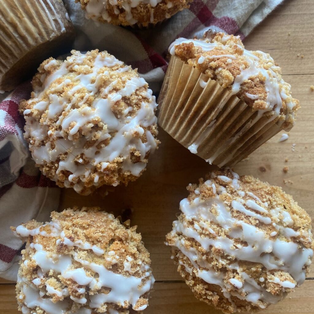 Freshly baked coffee cake muffins topped with buttery crumb streusel and a drizzle of sweet vanilla glaze, displayed on a wooden table with a rustic cloth