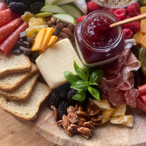 Holiday charcuterie board with assorted cheeses, cured meats, fresh fruit, nuts, toasted bread slices, and a jar of red raspberry jam.