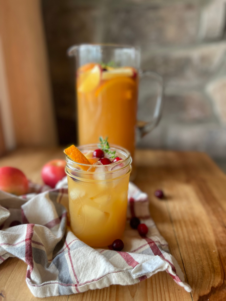 a large pitcher of apple cider punch is in the background, while a smaller glass full of punch is front and center. The glass is garnished with seasonal fruit like apples, cranberries, and orange slices