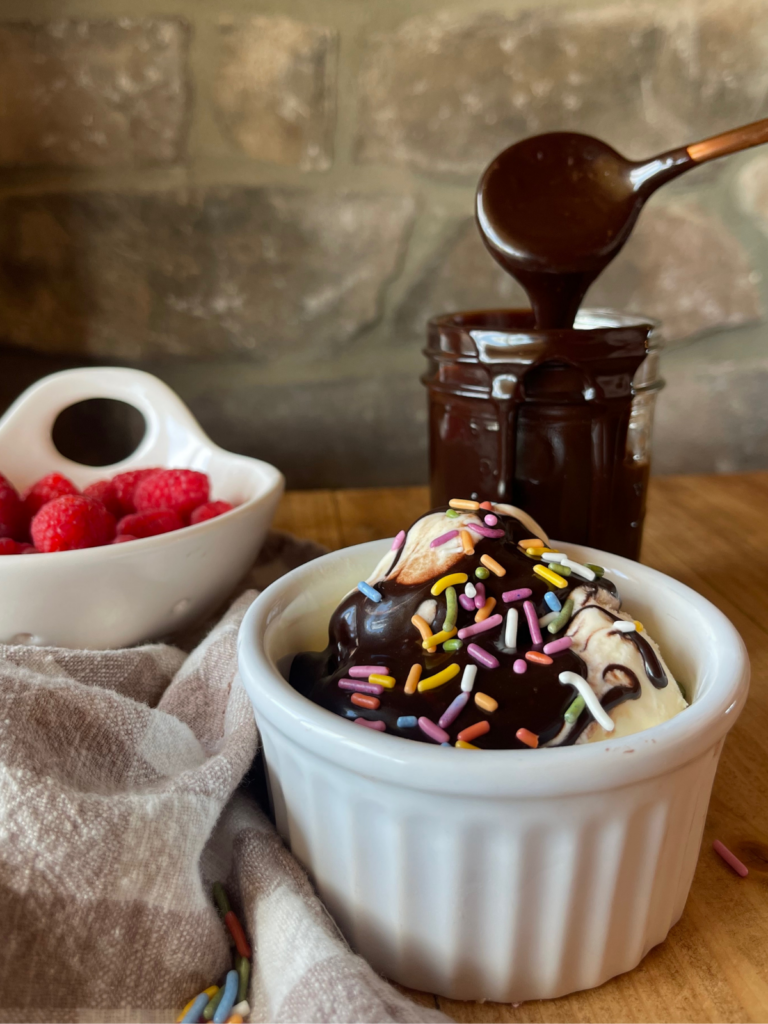 Homemade hot fudge sauce drizzled over a scoop of vanilla ice cream, which sits in a small white cup. A glass mason jar full of hot fudge is in the background