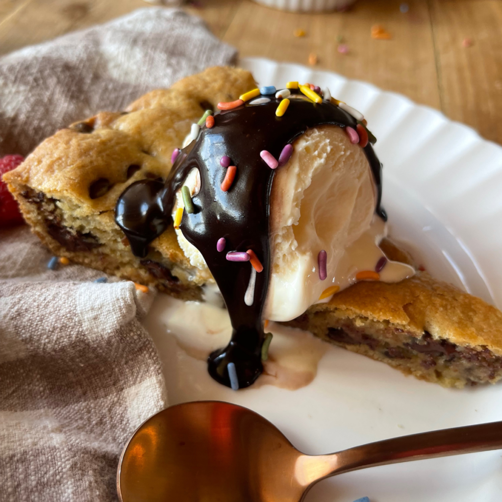a close up image of a slice of chocolate chip cookie cake topped with vanilla ice cream and a drizzle of homemade hot fudge