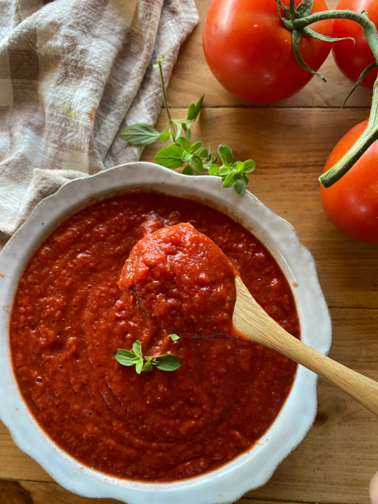 a bowl filled with homemade pizza sauce, garnished with fresh oregano. A wooden spoon is dipped slightly into the sauce