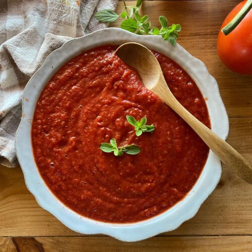 a bowl filled with homemade pizza sauce, garnished with fresh oregano. A wooden spoon is dipped slightly into the sauce