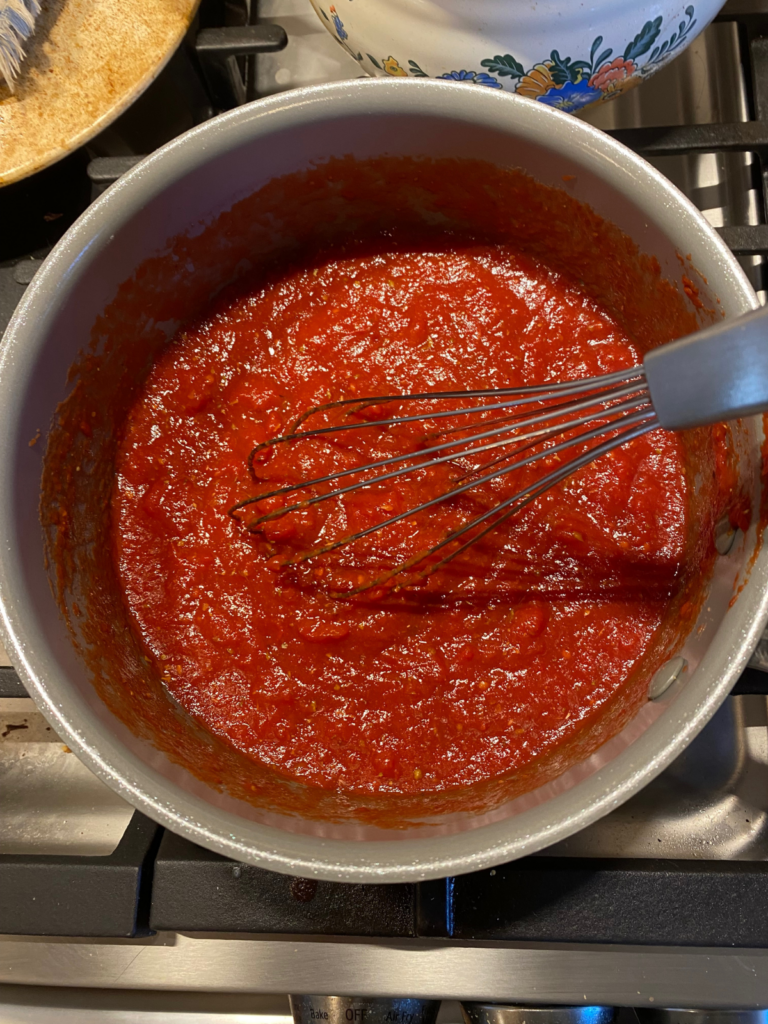 homemade pizza sauce in a saucepan on the stove. A whisk is in the middle of the pot, stirring the sauce
