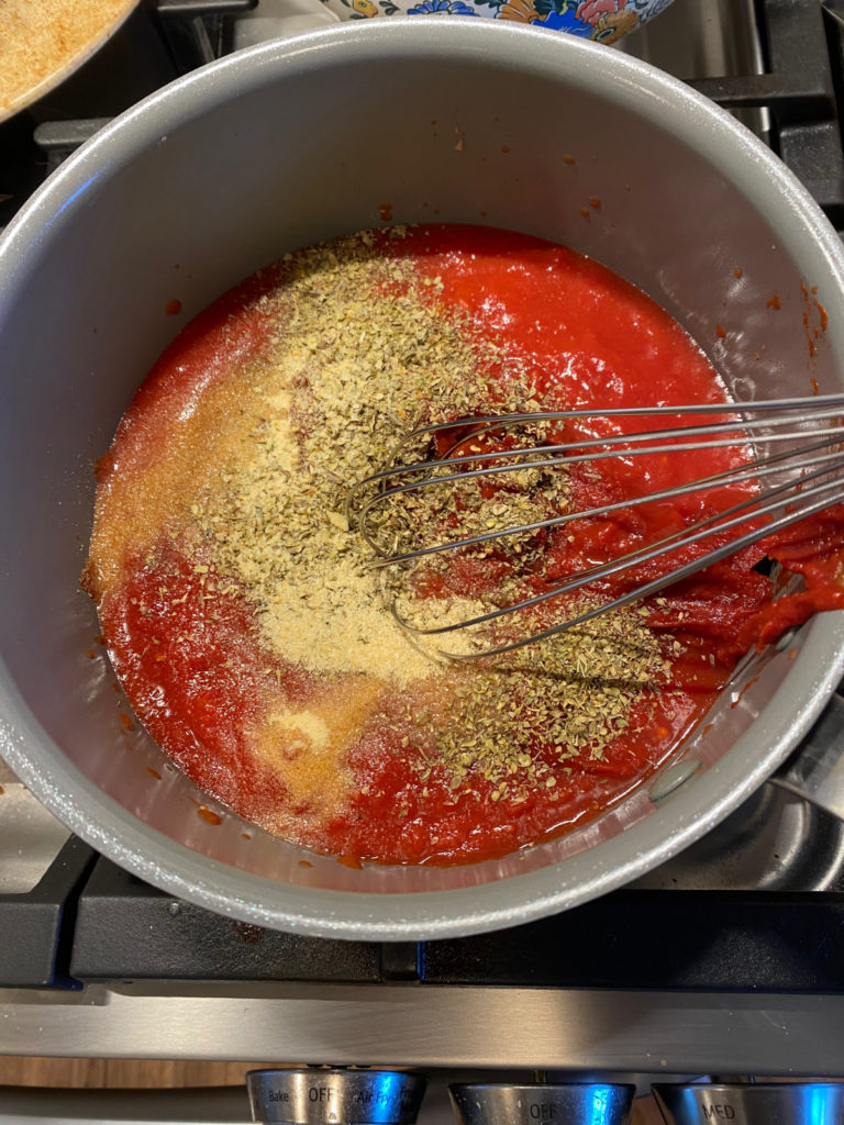 homemade pizza sauce in a saucepan on the stove. A whisk is in the middle of the pot, stirring the ingredients together